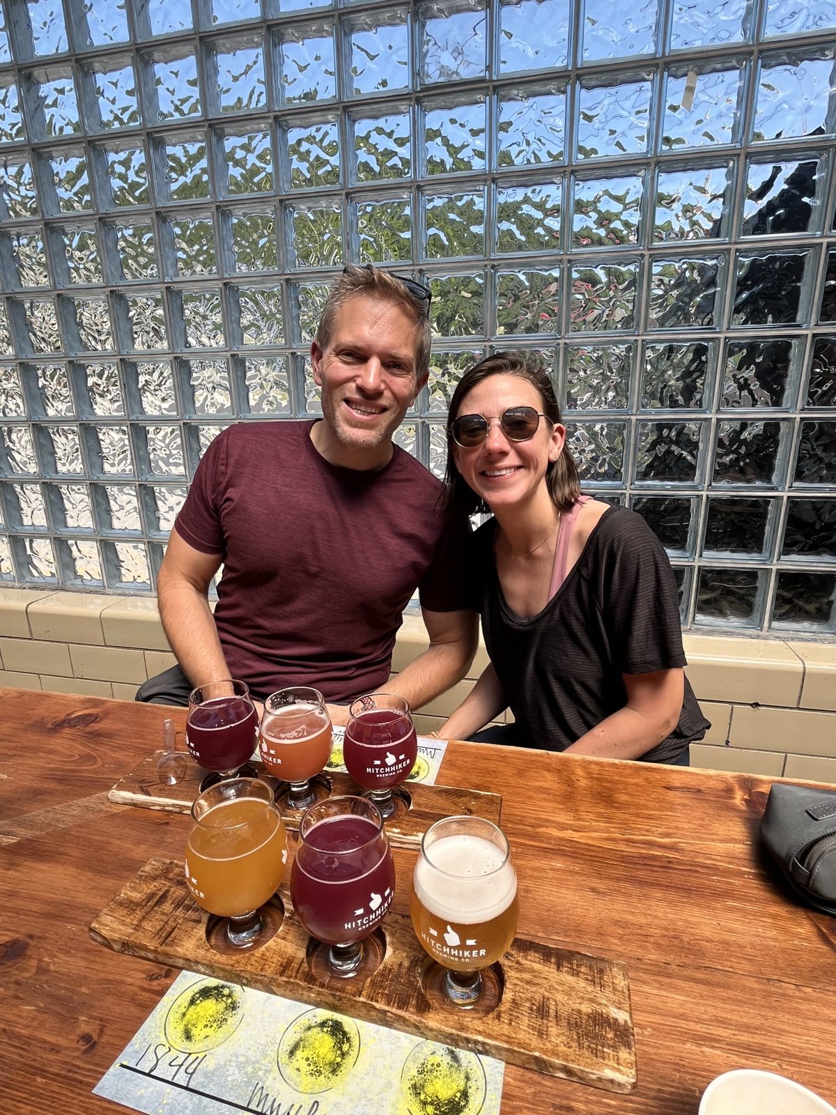 Haley and Michael behind two flights of beer at Hitchhiker Brewing Co., a glass-block wall behind them