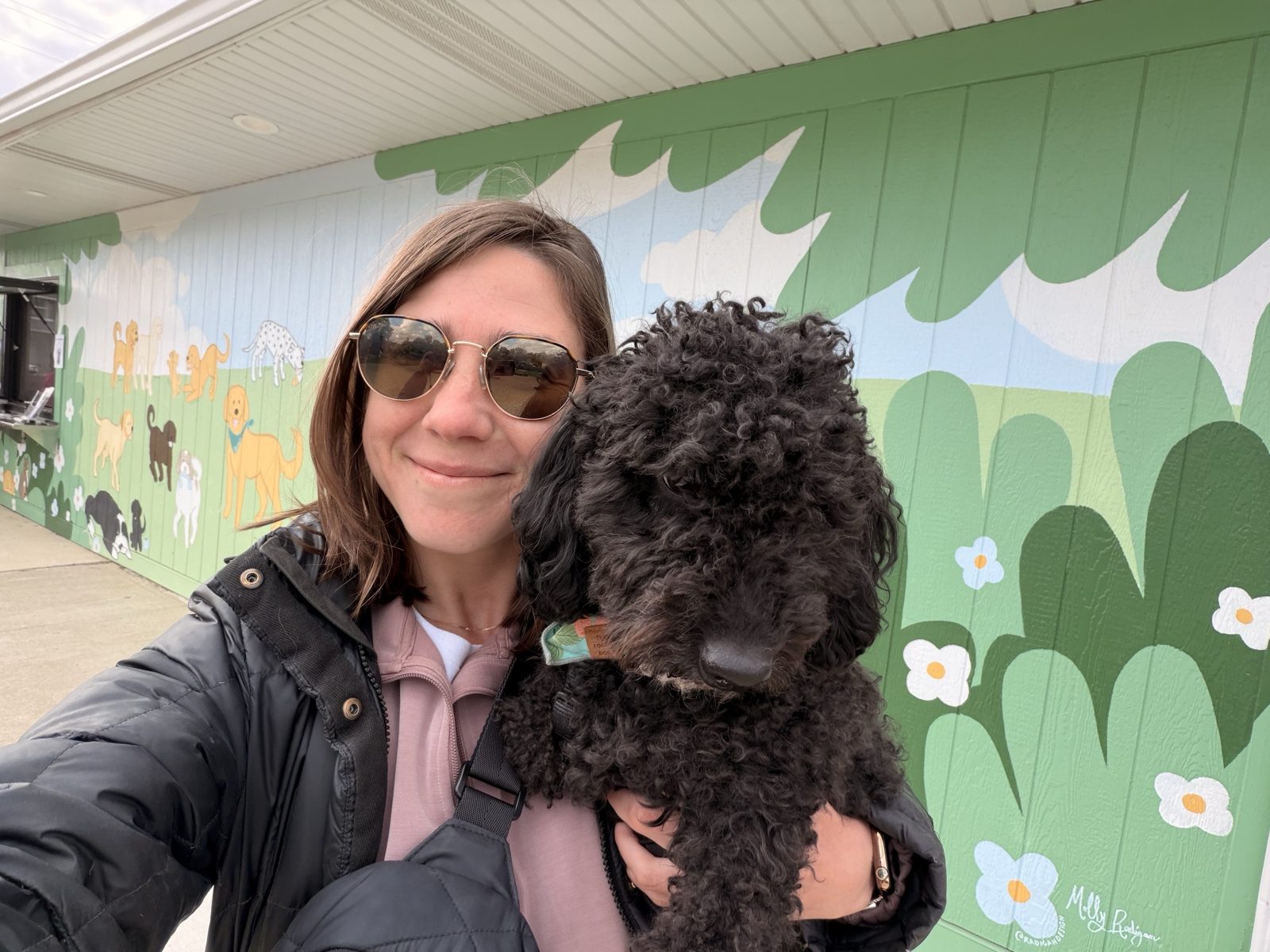 Haley in sunglasses taking a selfie holding Wes in front of a painted mural of dogs and flowers