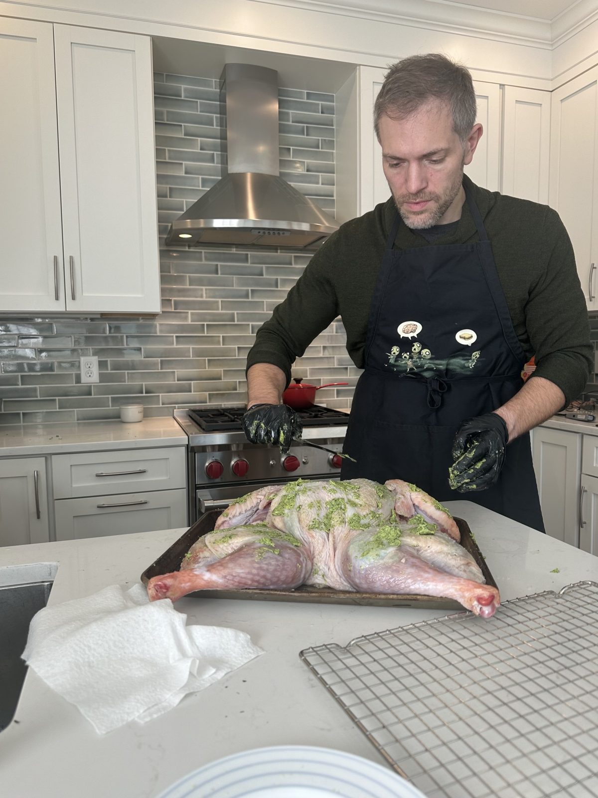 Michael in an apron and gloves rubbing green herb butter over a flattened turkey on a sheet pan