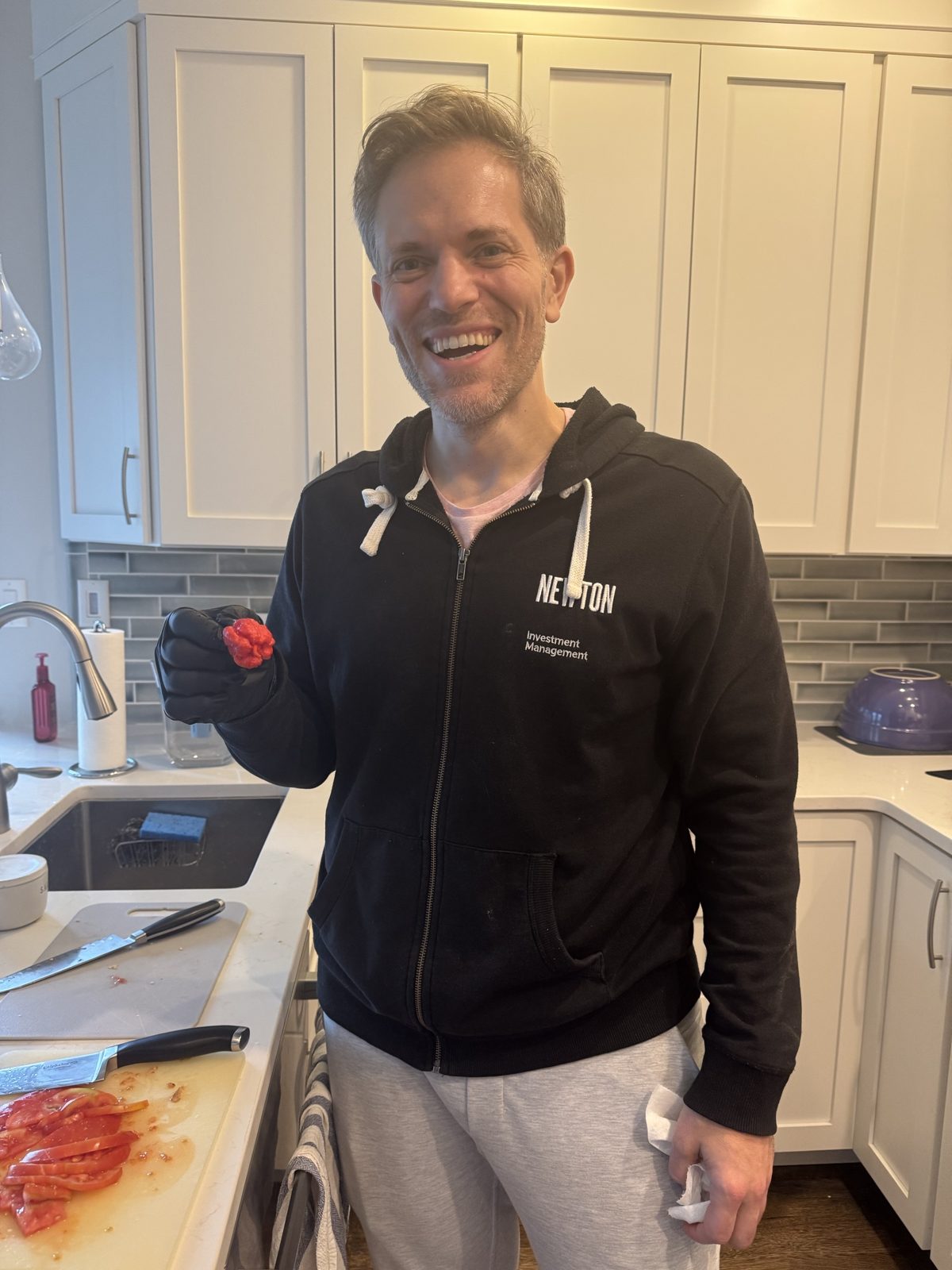 Michael in the kitchen grinning, holding a wrinkled red hot pepper in a gloved hand