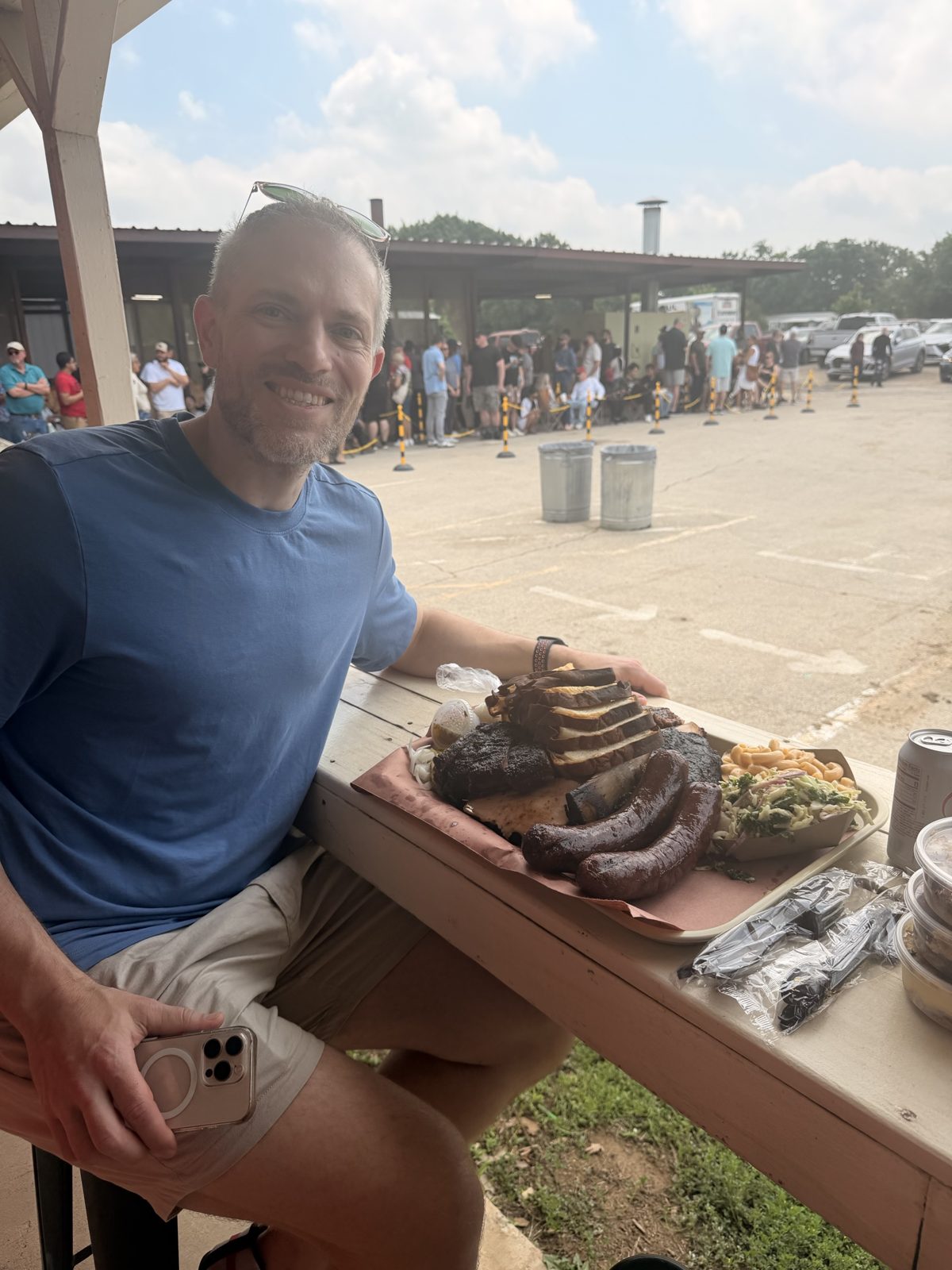 Michael at an outdoor picnic table with a tray of barbecue, sausages, and sides, a long line behind him