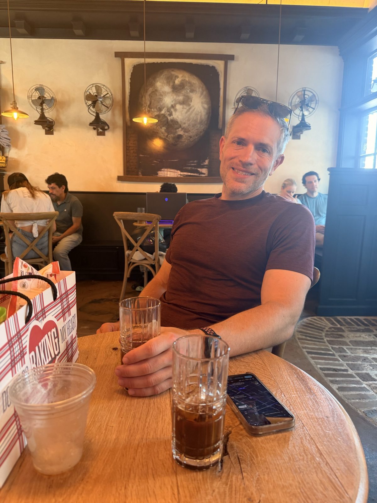 Michael at a round wooden cafe table with iced drinks, a large moon painting on the wall behind him