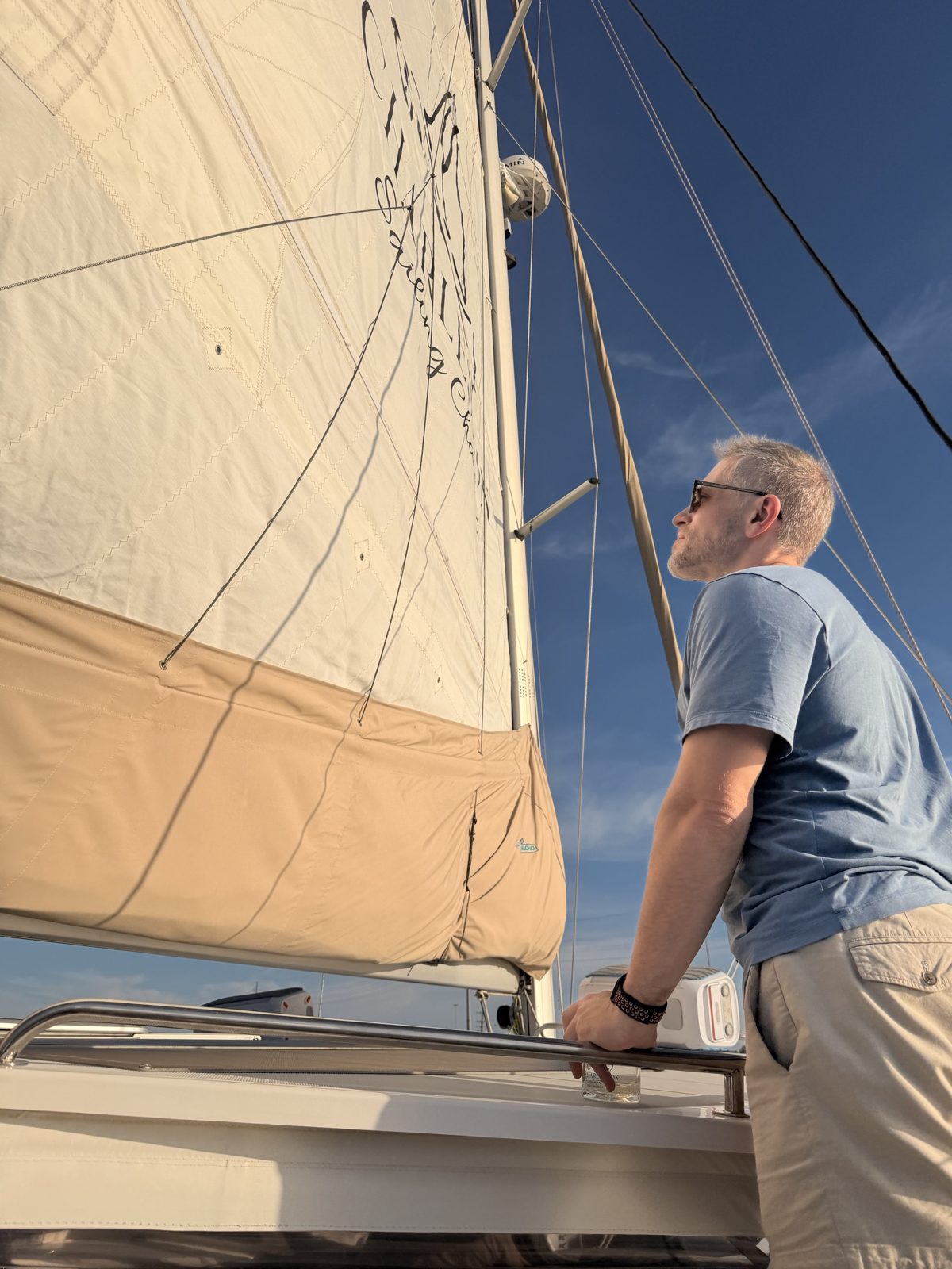 Michael on a sailboat deck, looking up at the mainsail against a blue sky